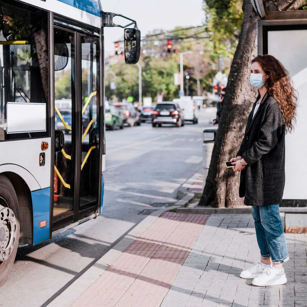 Junge Frau mit medizinischer Maske wartet an einer Bushaltestelle, während der Bus einfährt